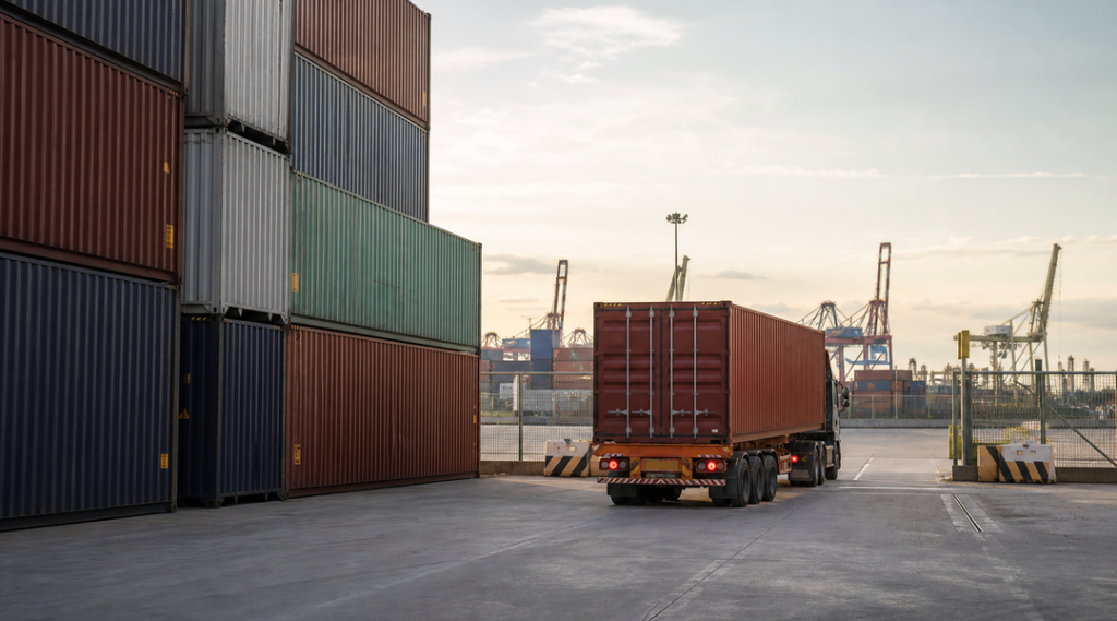 Loaded shipping containers stacked in a marine terminal yard beside a container chassis waiting near the terminal exit gate