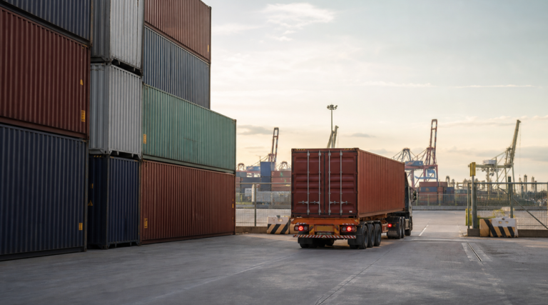 Loaded shipping containers stacked in a marine terminal yard beside a container chassis waiting near the terminal exit gate